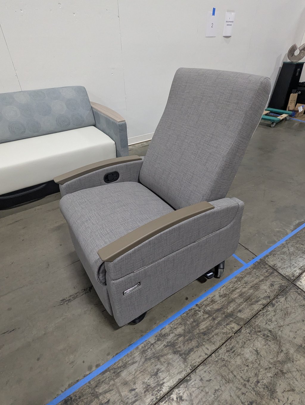 A grey recliner on casters beside a patterned bench at booth 2, floor marked with blue tape