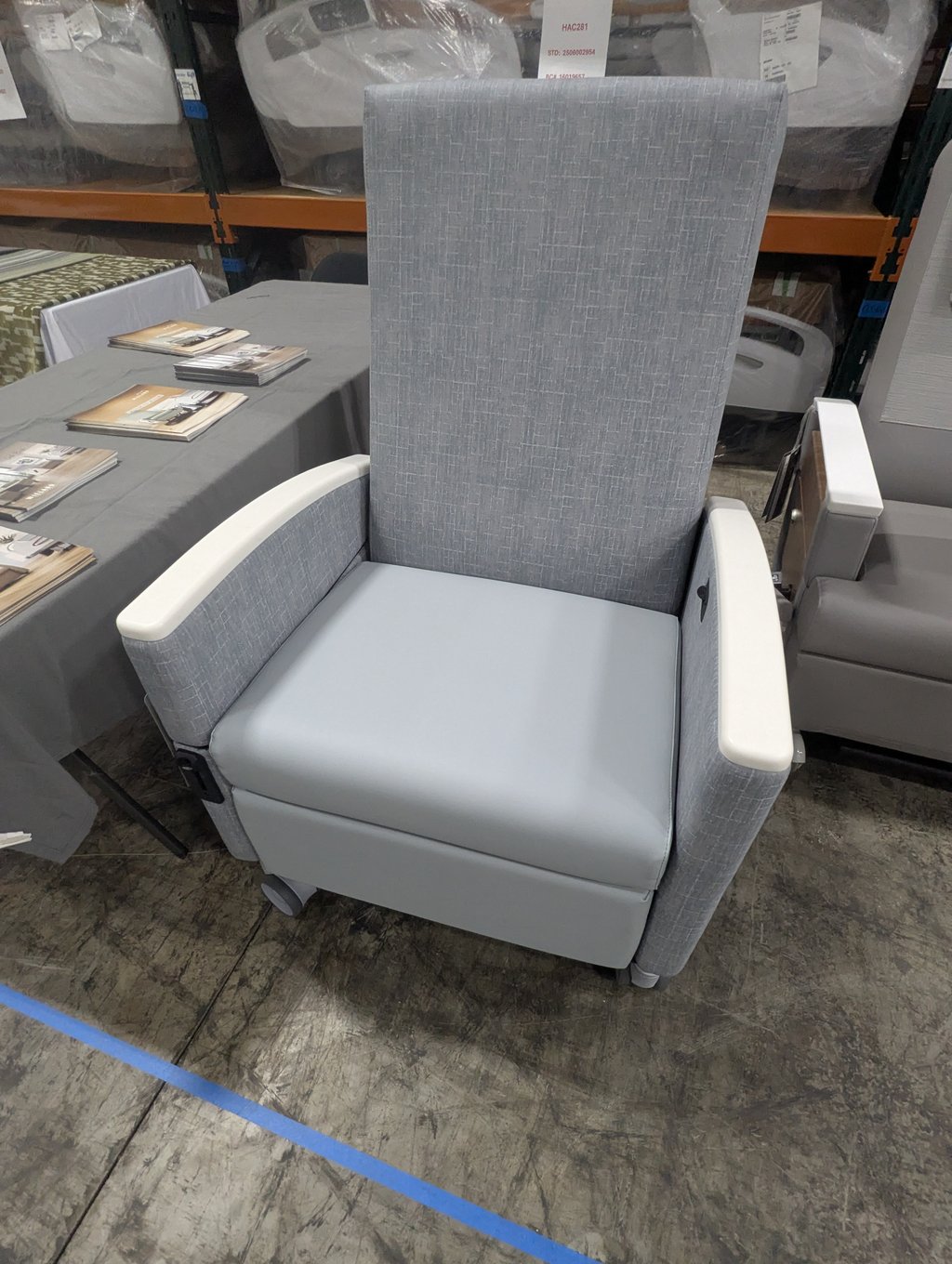 A tall light grey chair with white armrests beside a table of stacked brochures