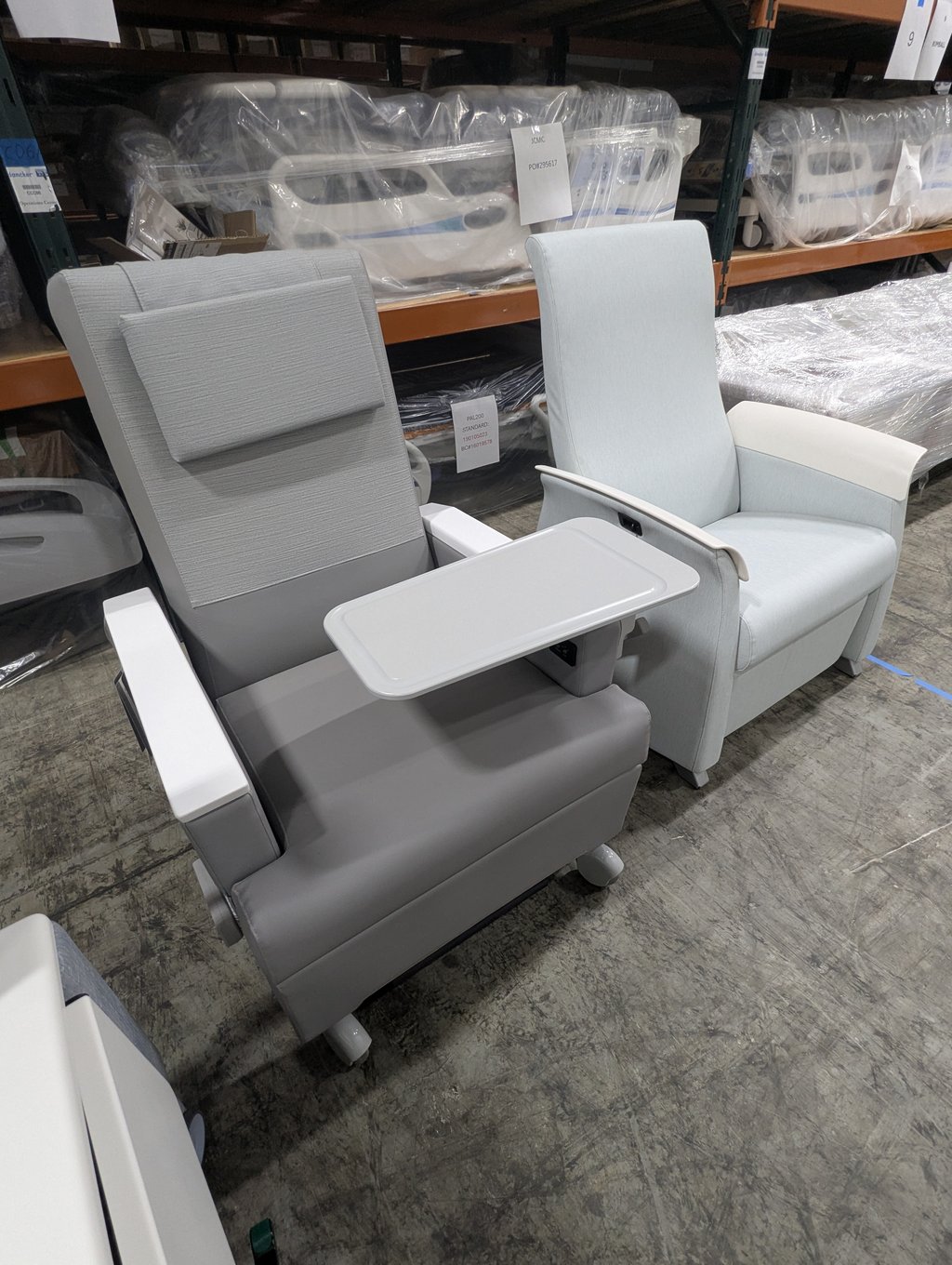 Two light grey medical recliners with fold-out trays in the warehouse aisle under a JCMC label