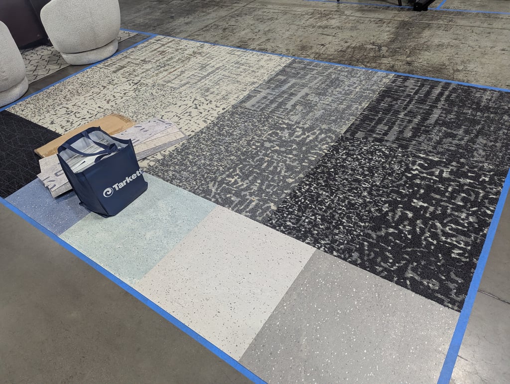 Carpet panels and vinyl tile samples taped out on the floor beside a Tarkett tote bag and two round stools