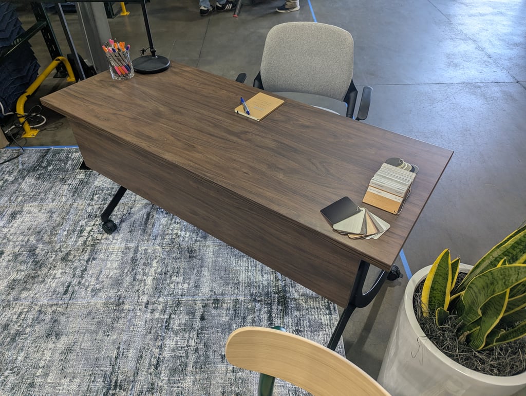 An overhead view of a dark wood desk with a grey chair, a jar of pens, a notebook and a fan of finish swatches