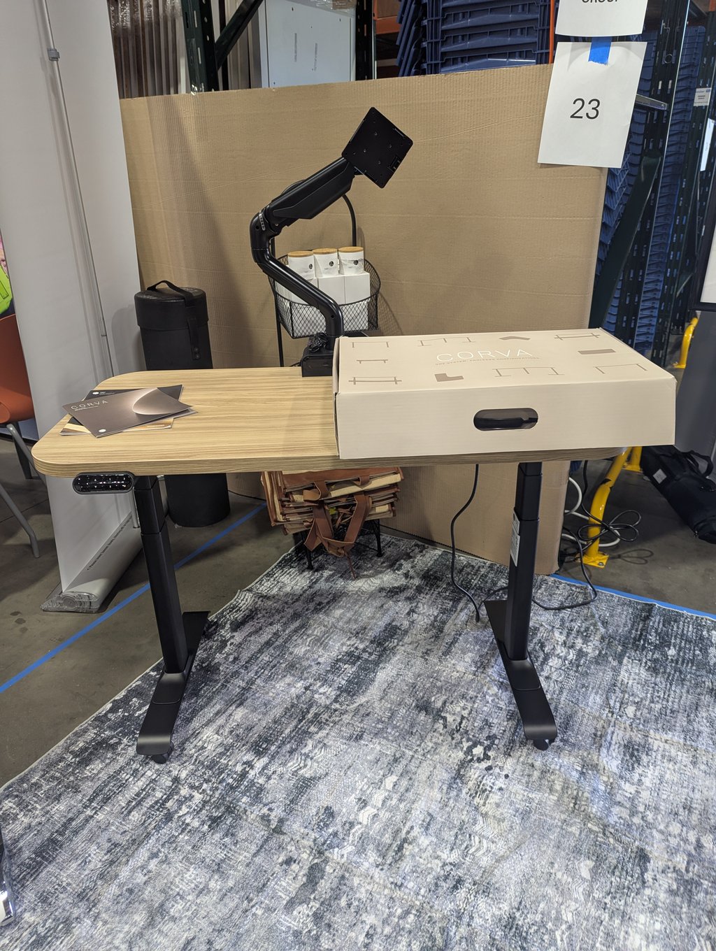 A wood-topped sit-stand desk with a monitor arm and a Corva box at booth 23