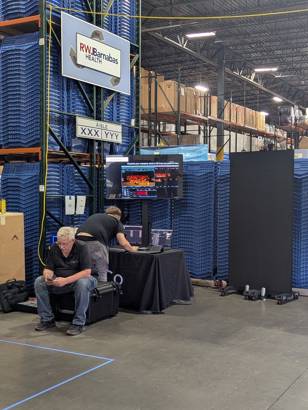 Two people working at laptops below the RWJBarnabas Health sign, with racks of blue bins behind