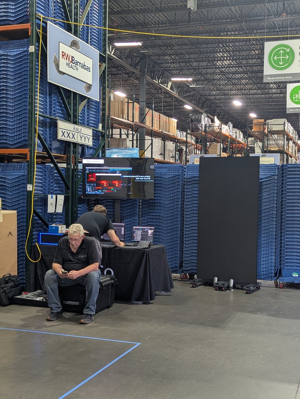 Warehouse racking filled with blue bins beside a table of monitors and laptops under the RWJBarnabas Health sign