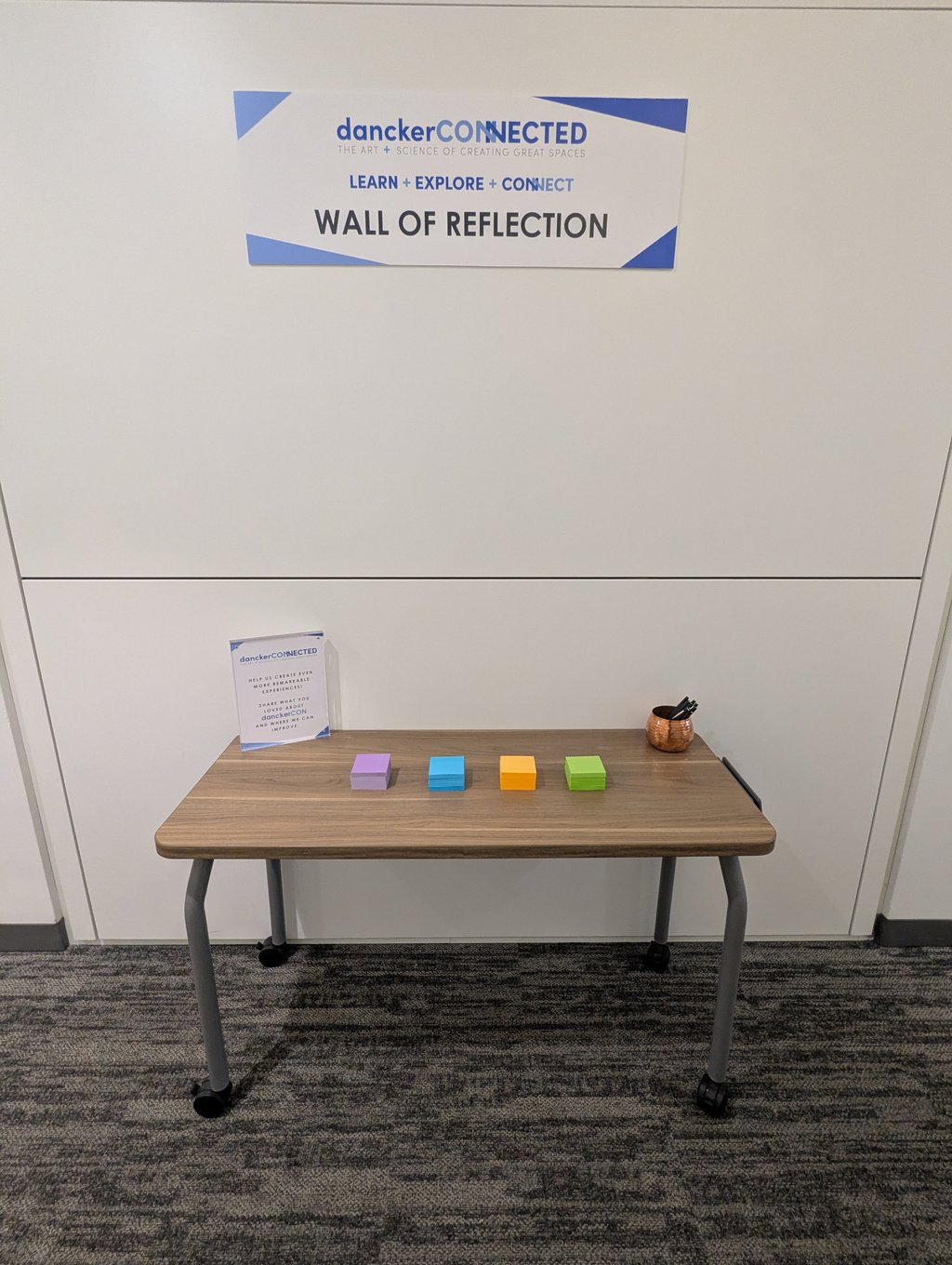 The danckerCONNECTED Wall of Reflection sign above a table of sticky note pads and a copper pen cup