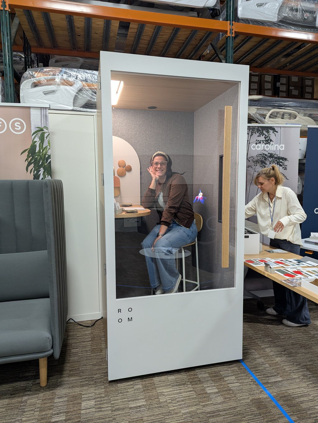 An attendee seated inside a white acoustic pod while another looks at colour samples beside the Carolina banner