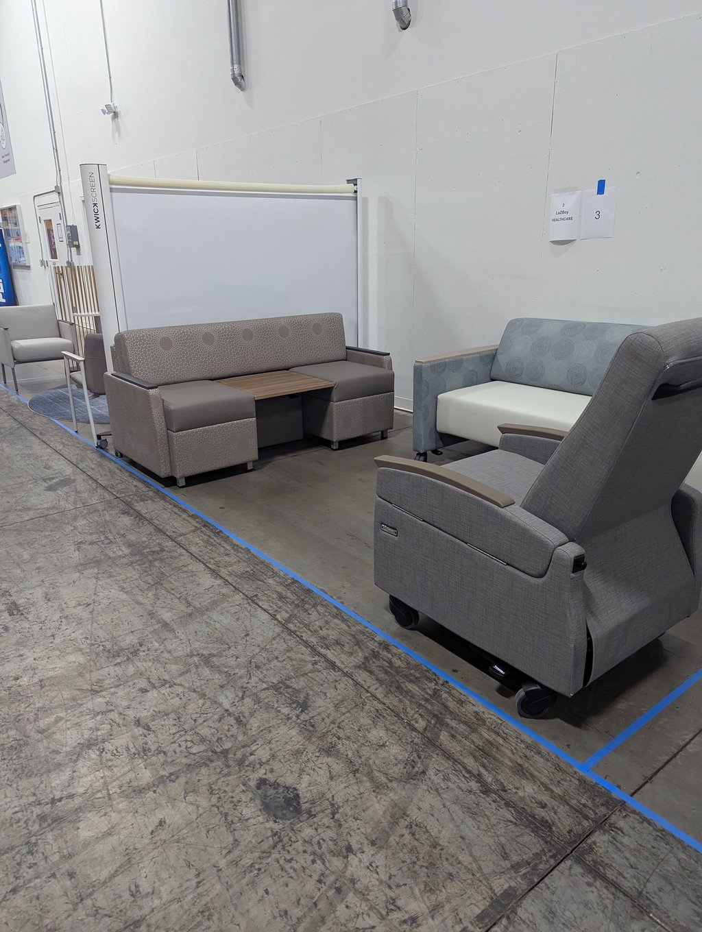 Grey healthcare lounge chairs and sofas beside a KwickScreen privacy screen at the La-Z-Boy Healthcare booth, number 3