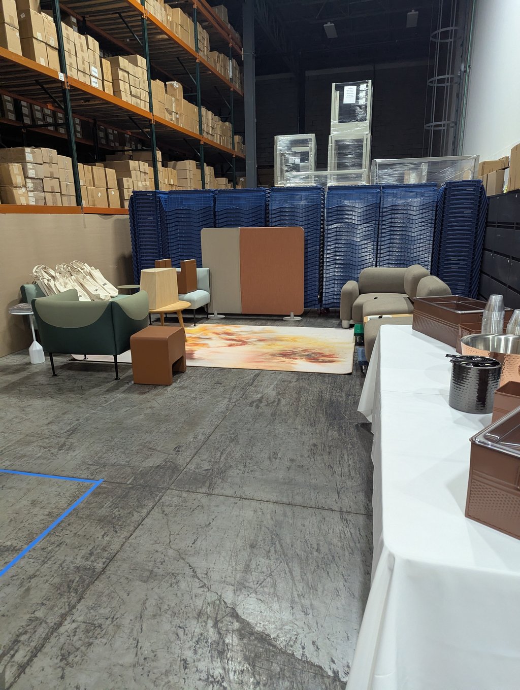 A lounge setup of green and beige chairs on a colourful rug beside a catering table, with warehouse racking behind