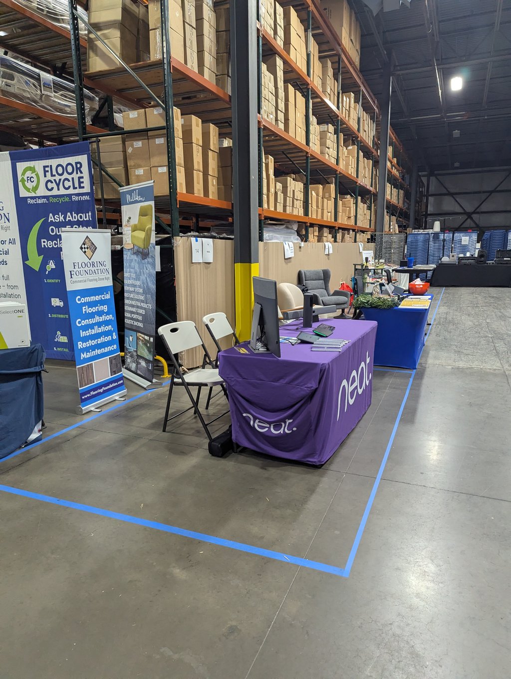 A row of exhibitor tables and chairs below tall pallet racks, with Floor Cycle and Flooring Foundation banners