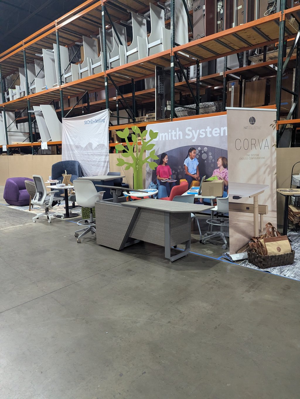 Classroom and office furniture with a green tree cutout between the Smith System, Corva and SitOnIt Seating stands