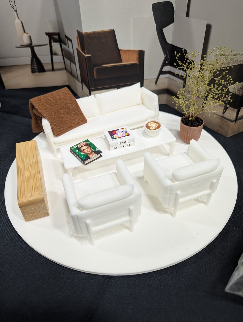 A white scale model of a living room with a sofa, armchairs, coffee table and potted plant on a round base