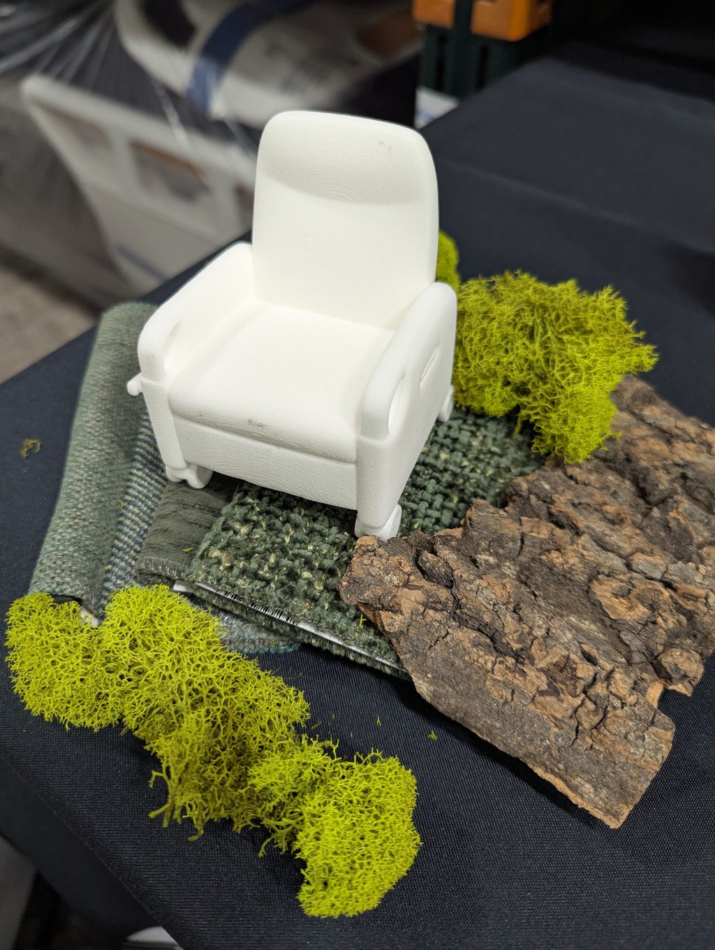 A small white model armchair resting on green textiles, bark and bright moss clumps