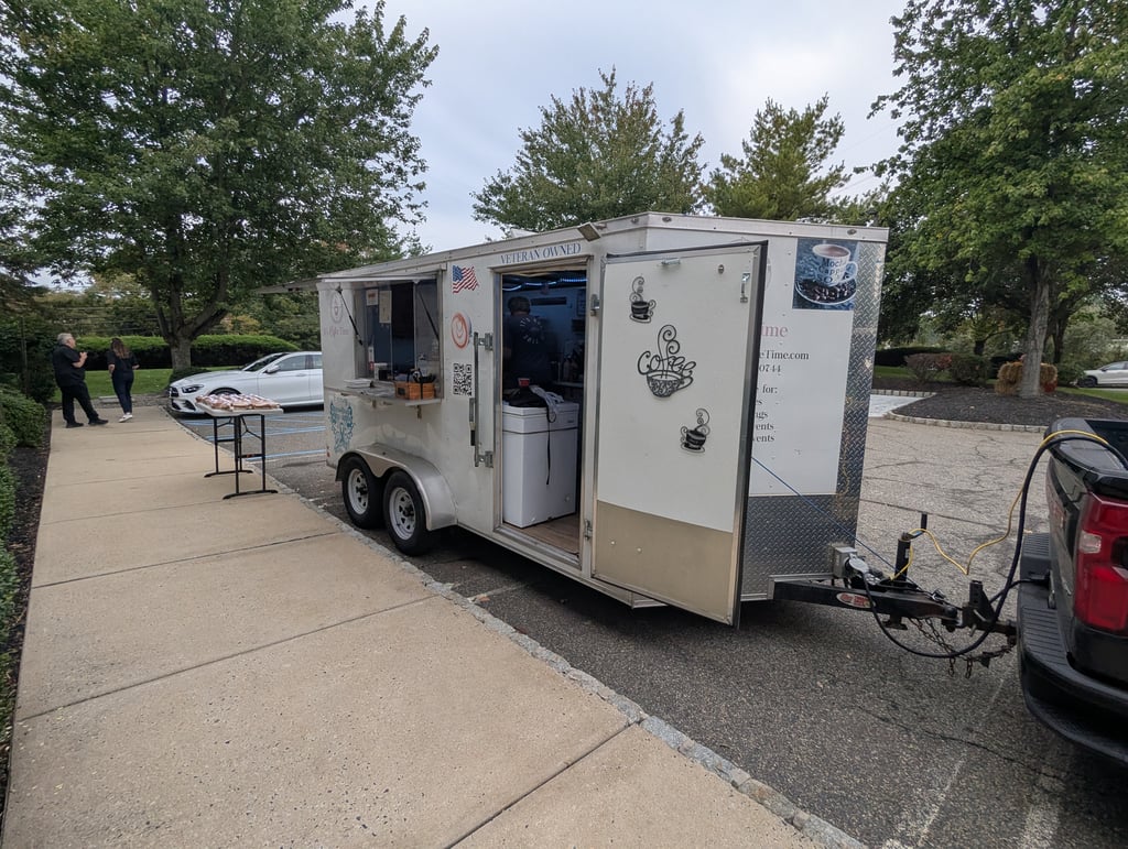 A white veteran-owned coffee trailer parked at the curb with its serving window open and a pastry table beside it