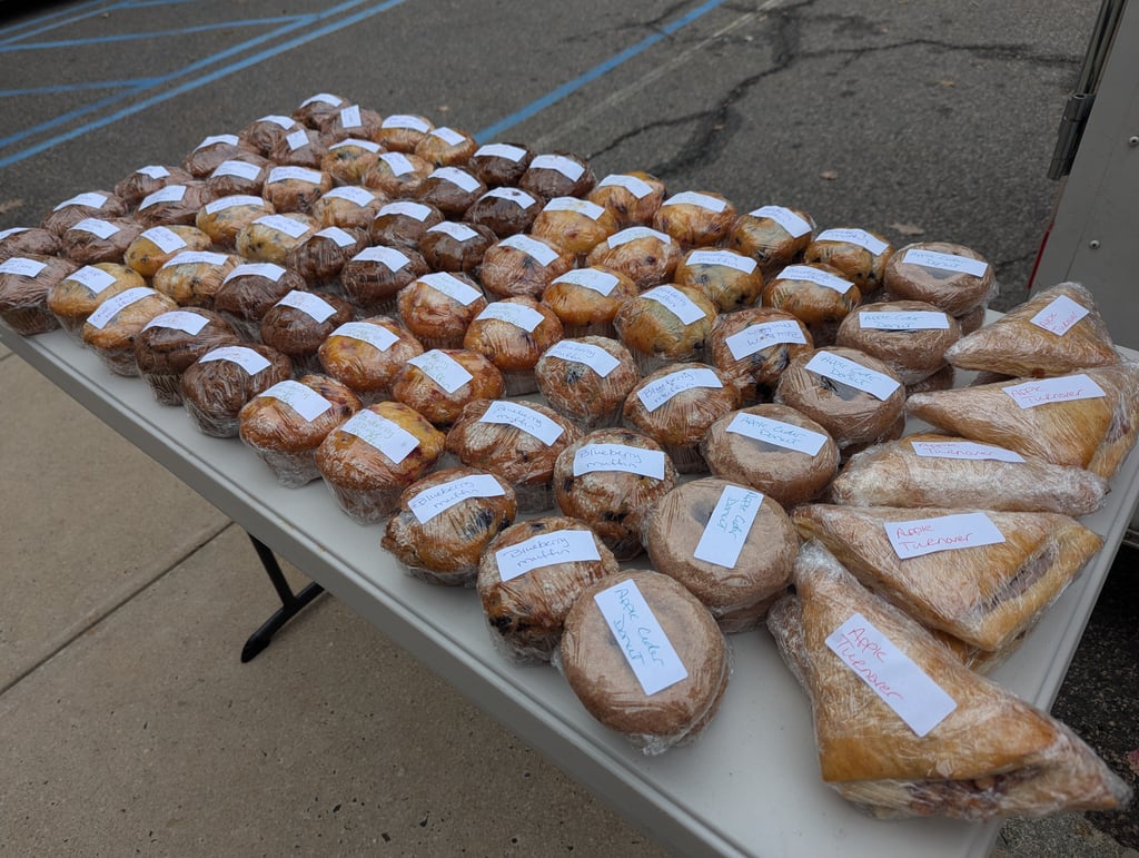 Rows of wrapped muffins, donuts and turnovers on a folding table, each with a handwritten label