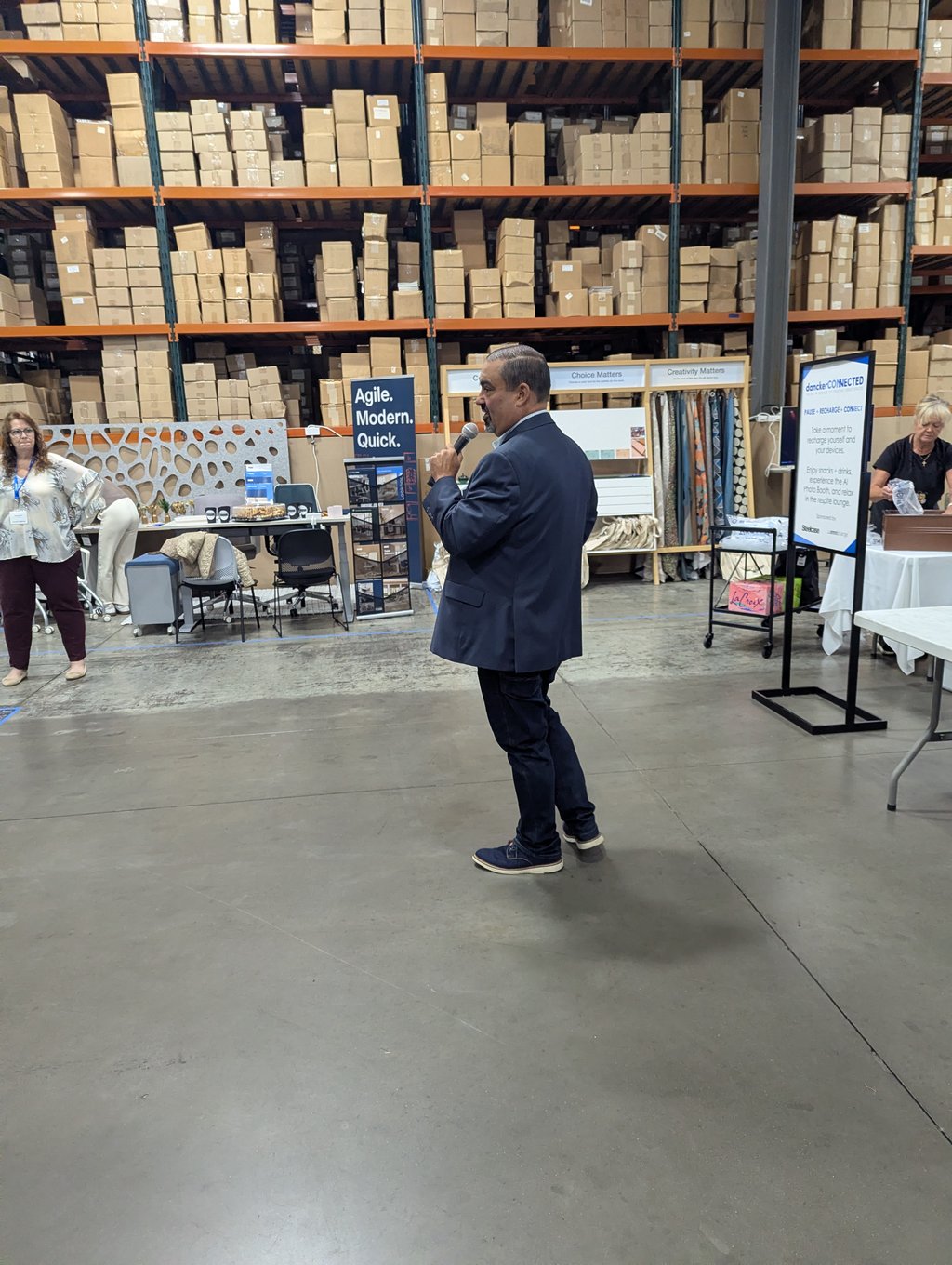 A speaker in a suit holding a microphone on the event floor, with booths and attendees along the racking behind
