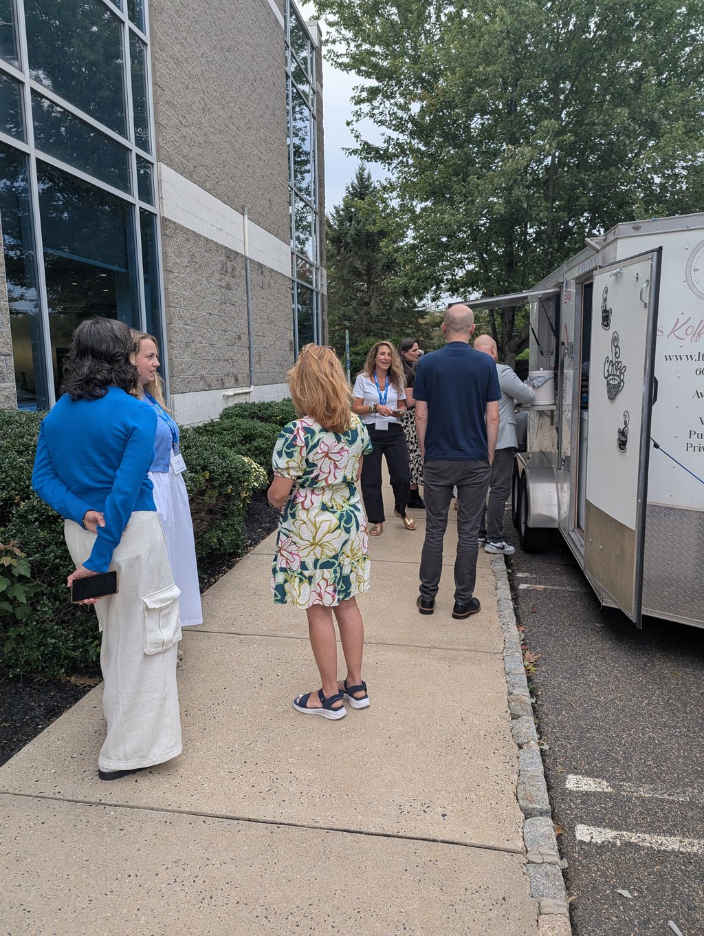 Attendees chatting in line on the sidewalk beside the coffee trailer and the office building