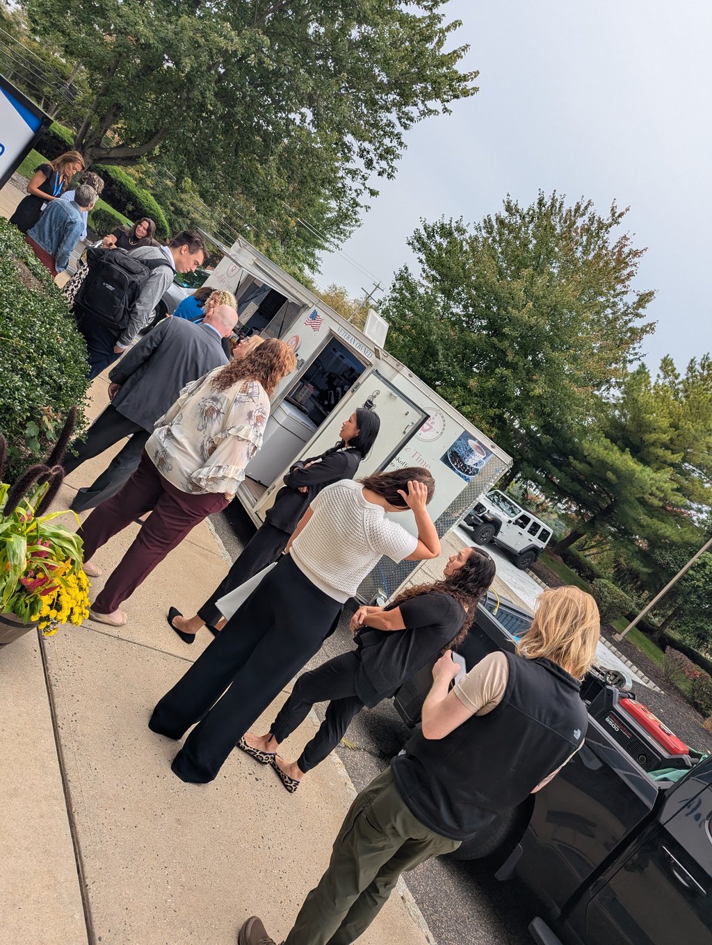 Attendees queuing on the sidewalk at the white veteran-owned food trailer, with trees and a flower planter behind