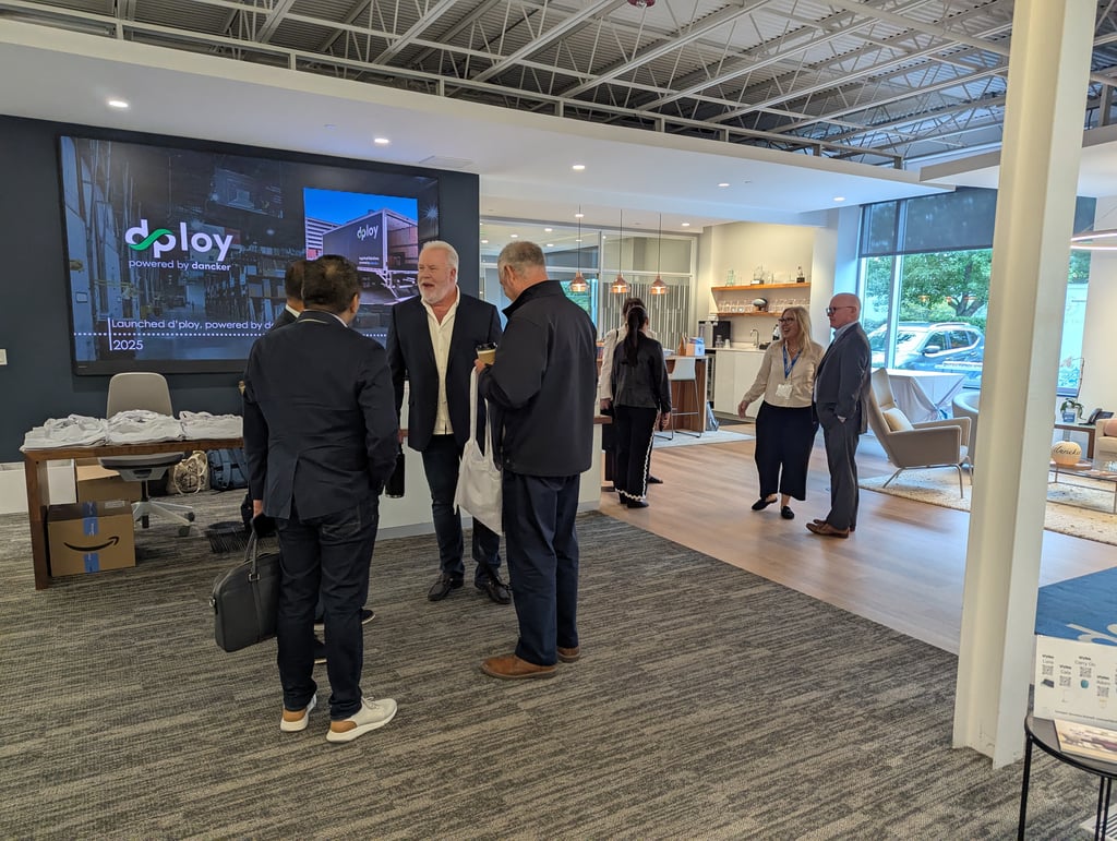 People talking in an office lounge beside a large wall screen showing the dploy logo and powered by dancker
