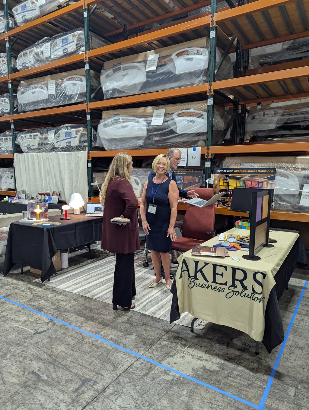 Three people talking at the Akers Business Solutions booth beneath pallet racking of wrapped components