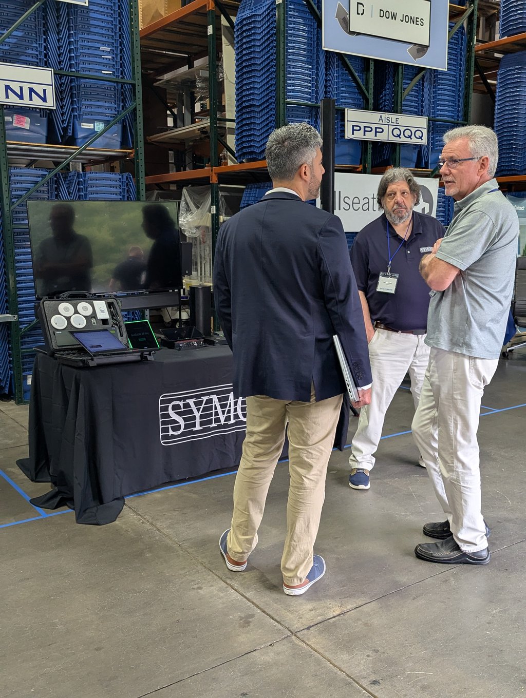 Three attendees talking at a booth of monitors and equipment beside stacked blue bins under the Dow Jones sign