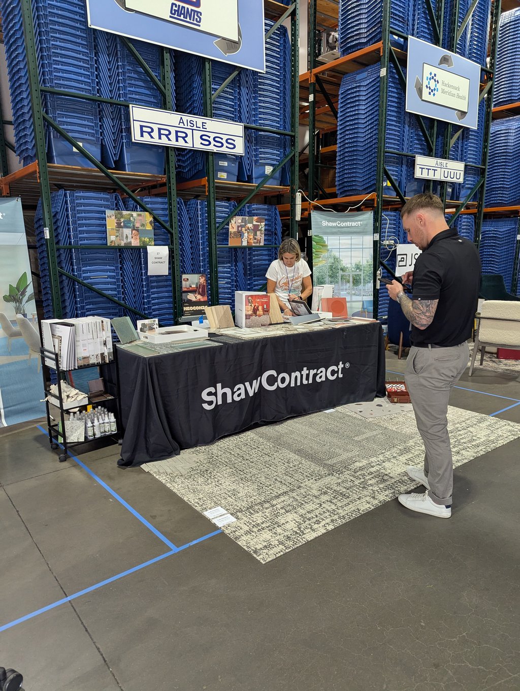 Sample displays on the ShawContract booth table with racks of blue bins behind and two exhibitors at the stand