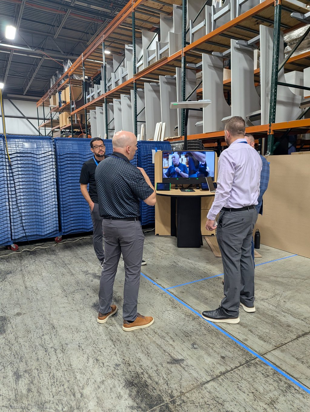 Four attendees gathered around a demo kiosk showing a live video feed, with orange racking behind