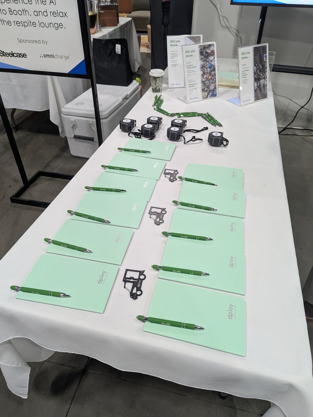 Rows of mint green dploy notebooks, pens and tape measures on a giveaway table with information cards