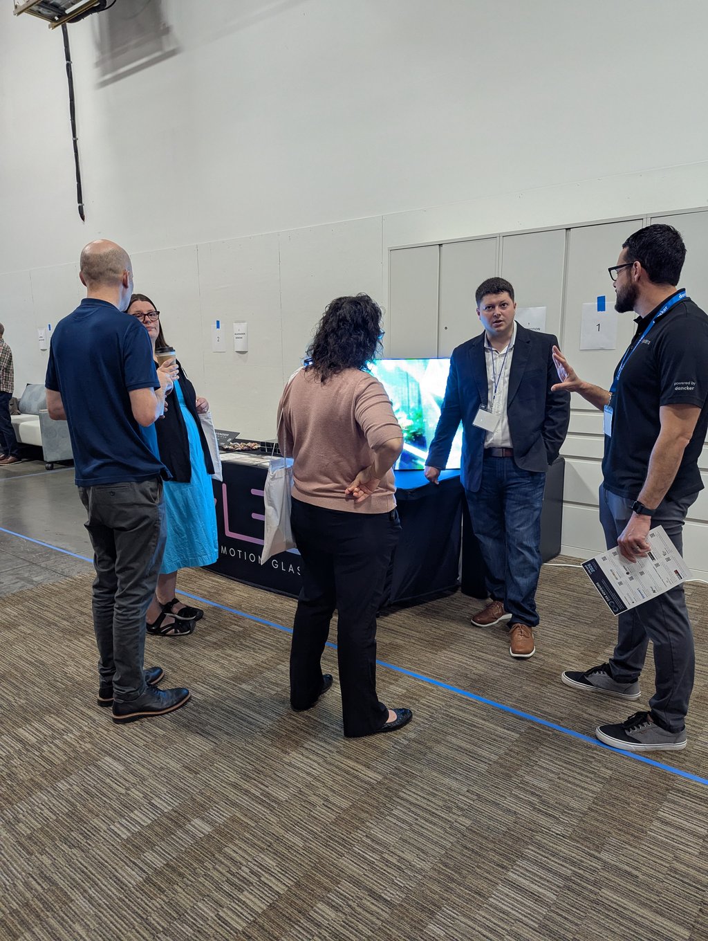 Five attendees talking and holding brochures at the Clear Motion Glass booth with its bright display
