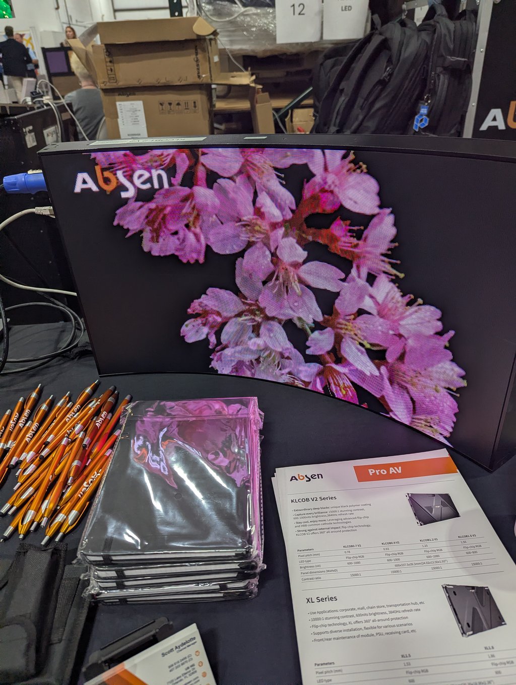 A curved Absen LED display showing large pink flowers above brochures and orange pens on the table