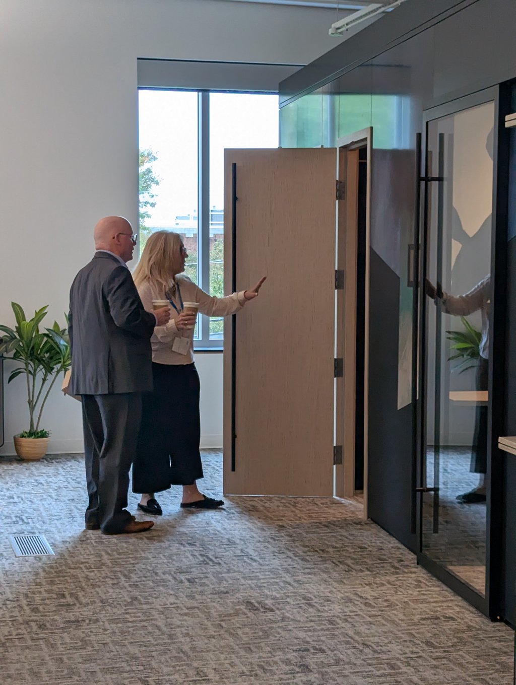 Two attendees with takeaway coffee talking in a carpeted office corridor beside a glass partition