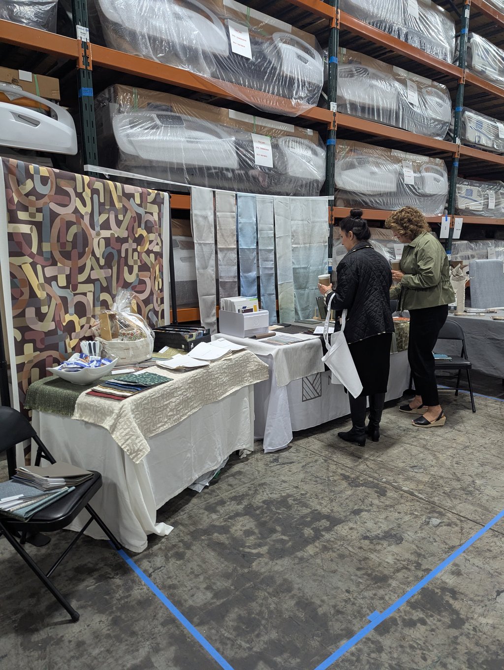 Two attendees examining fabric swatches and sample books at a display table in the warehouse aisle