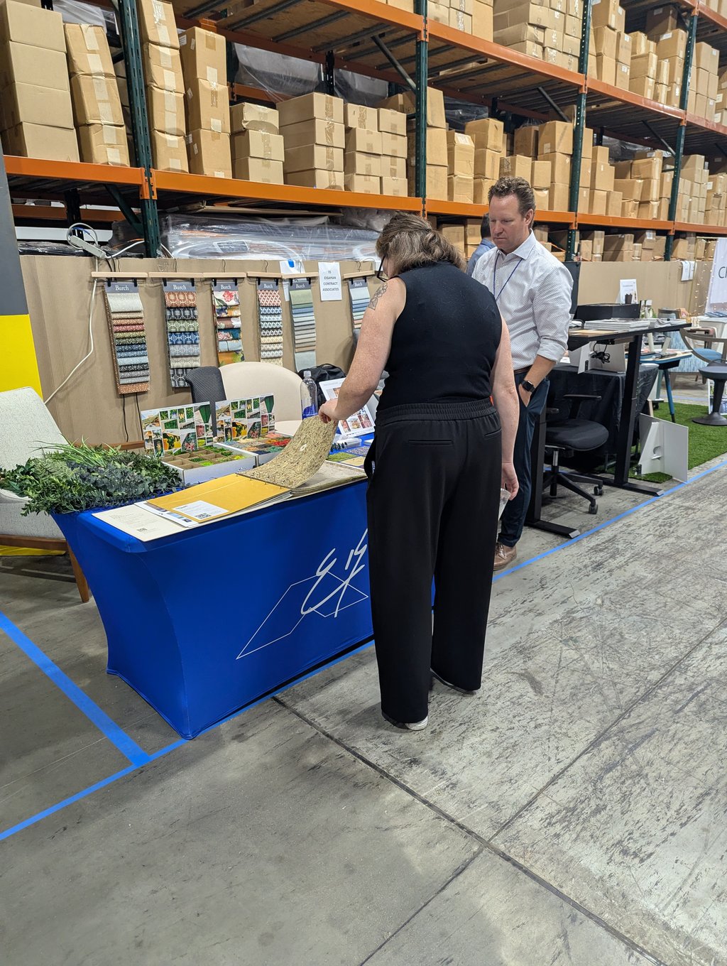 Two attendees examining Burch fabric and flooring samples at a table in the warehouse aisle