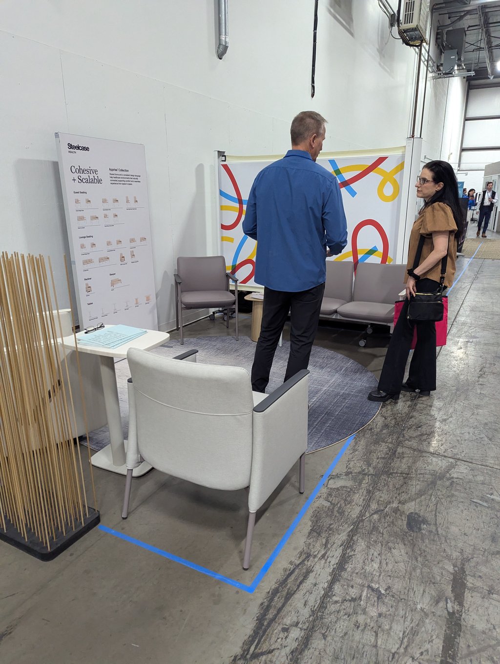 Two attendees talking among grey lounge chairs beside the Steelcase Cohesive and Scalable display board