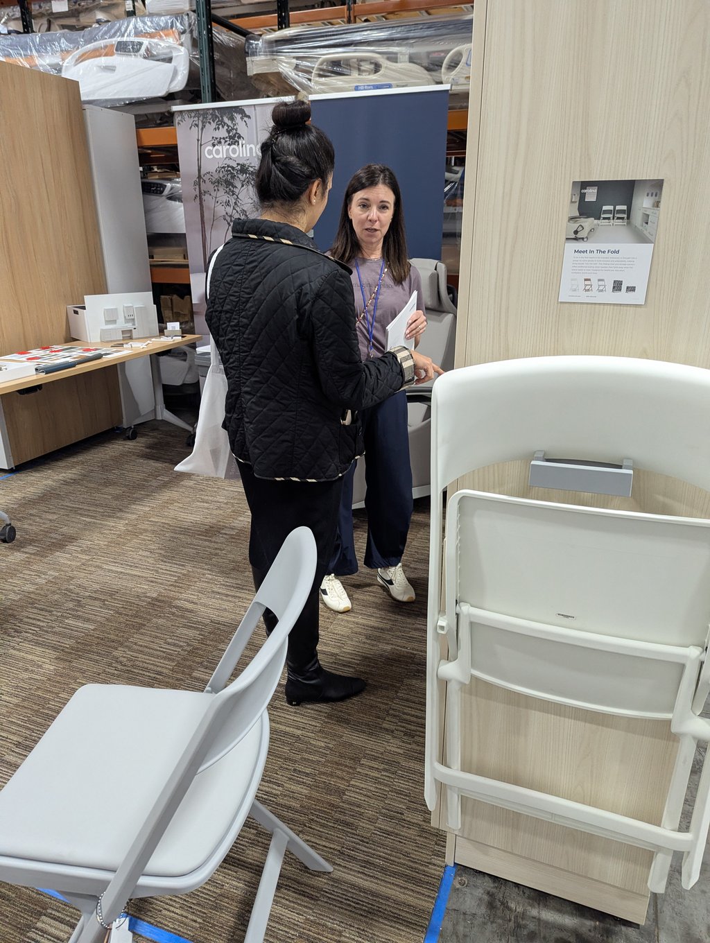 Two attendees talking among stacked folding chairs beside the Carolina banner and Meet In The Fold poster