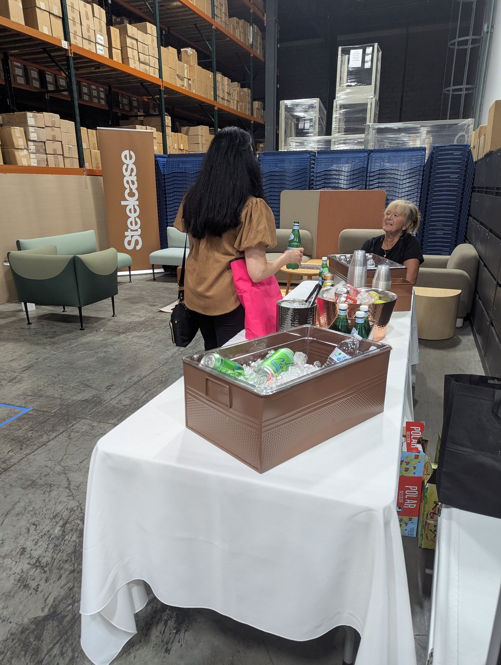 Two attendees talking beside a table of iced drinks under the Steelcase banner in the warehouse lounge