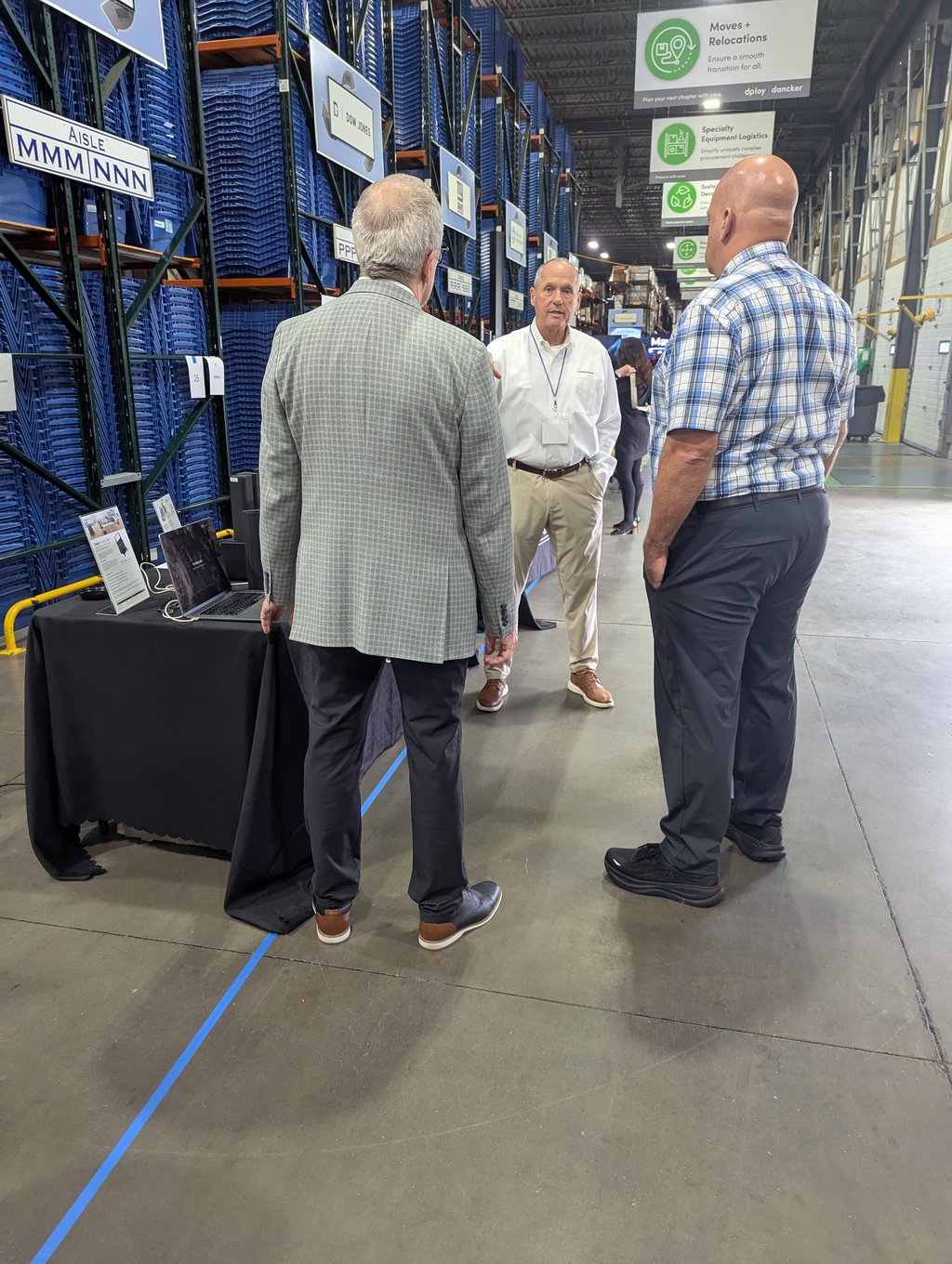 Three attendees talking in an aisle of blue bin racking beneath the Moves and Relocations sign