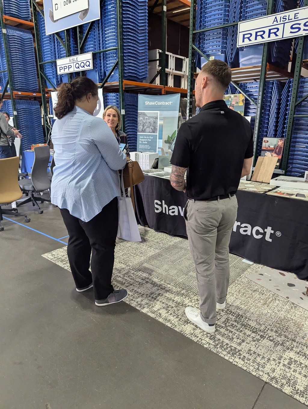 Three attendees at the ShawContract stand with racks of blue bins and aisle signage behind