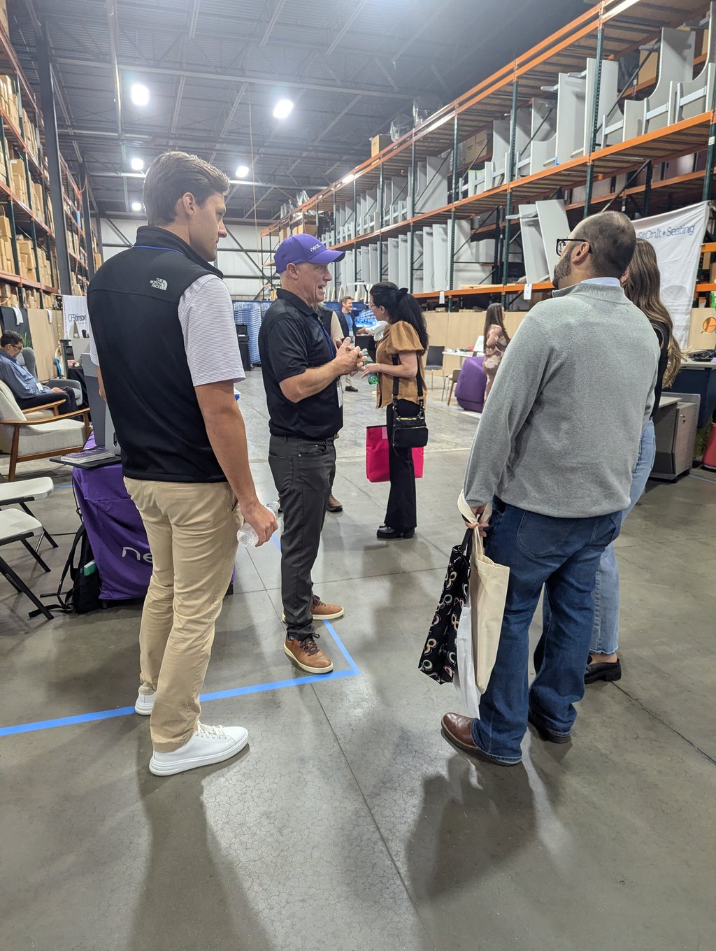 Attendees standing and talking in a wide aisle lined with tall orange shelving and stacked furniture