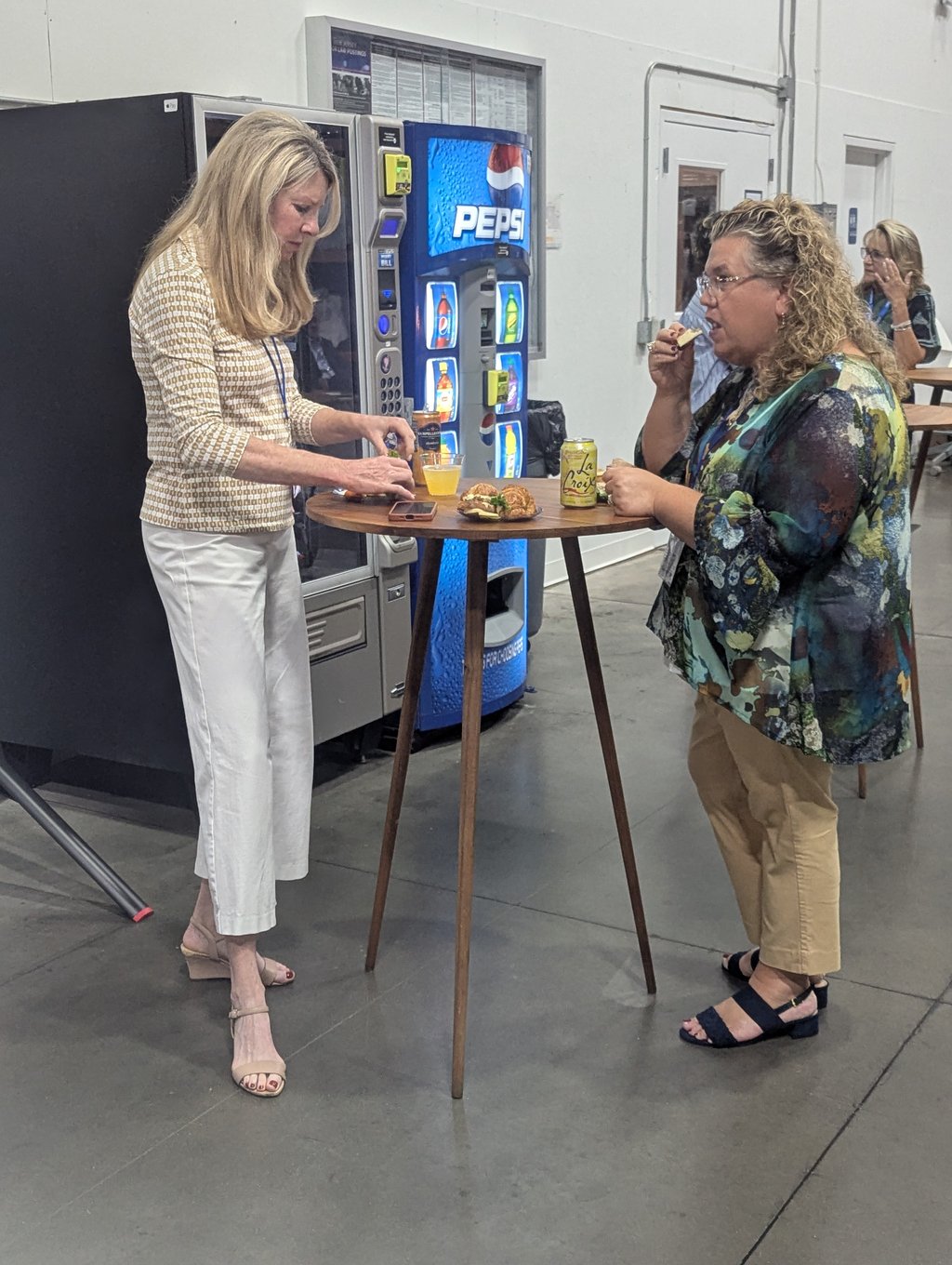 Two attendees eating at a tall round table with drinks and small sandwiches, beside a vending machine