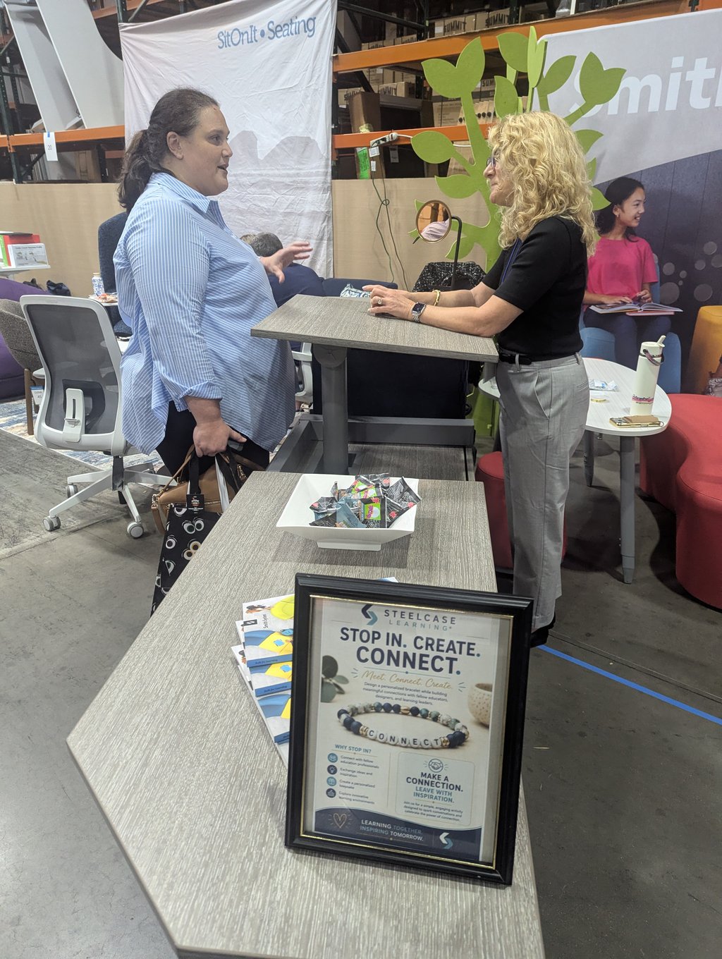Two attendees talking across a raised demo table under the SitOnIt Seating banner and a Stop In Create Connect sign