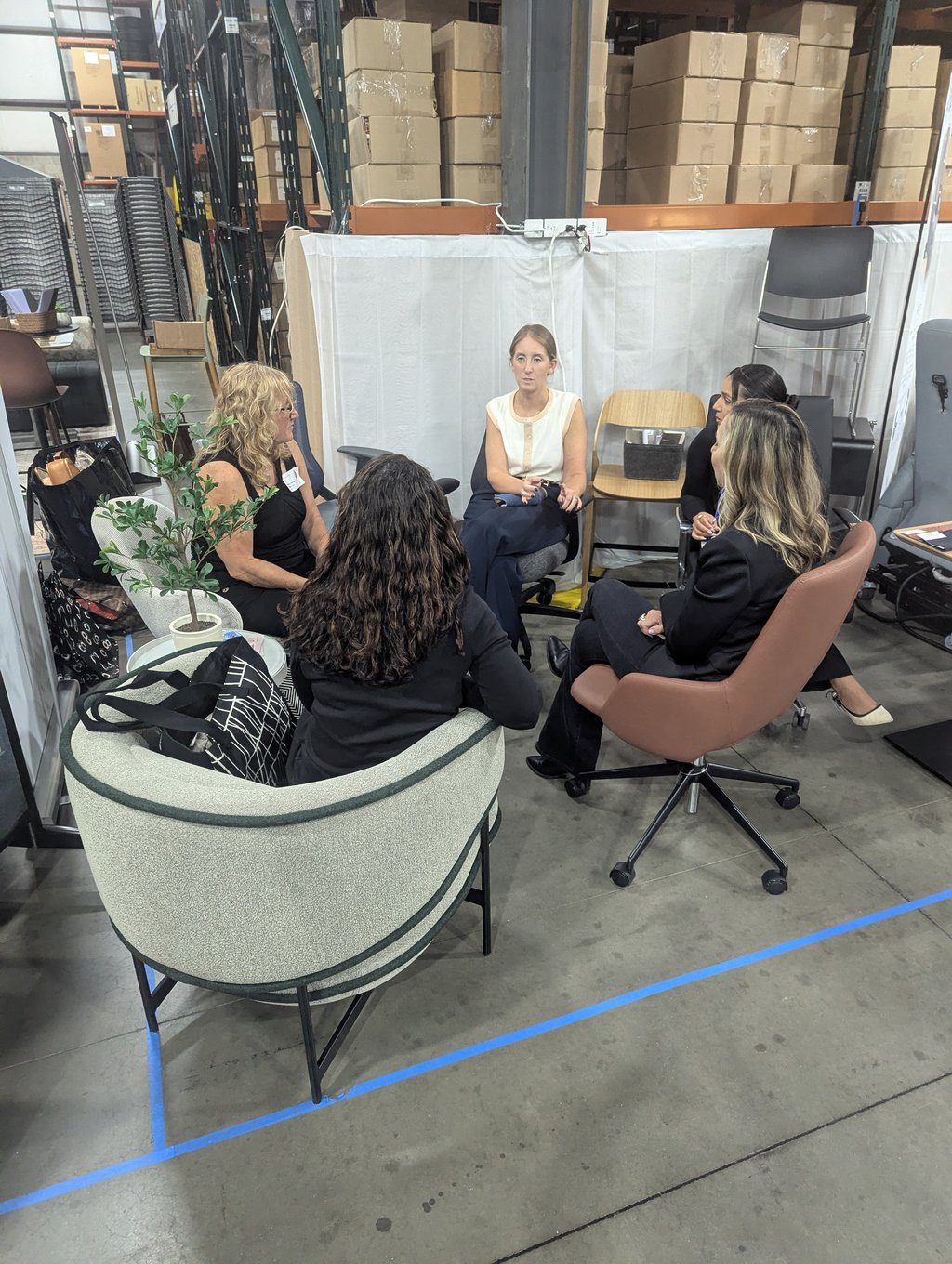 Five attendees seated in a circle of office chairs in discussion, with racking and a white partition behind
