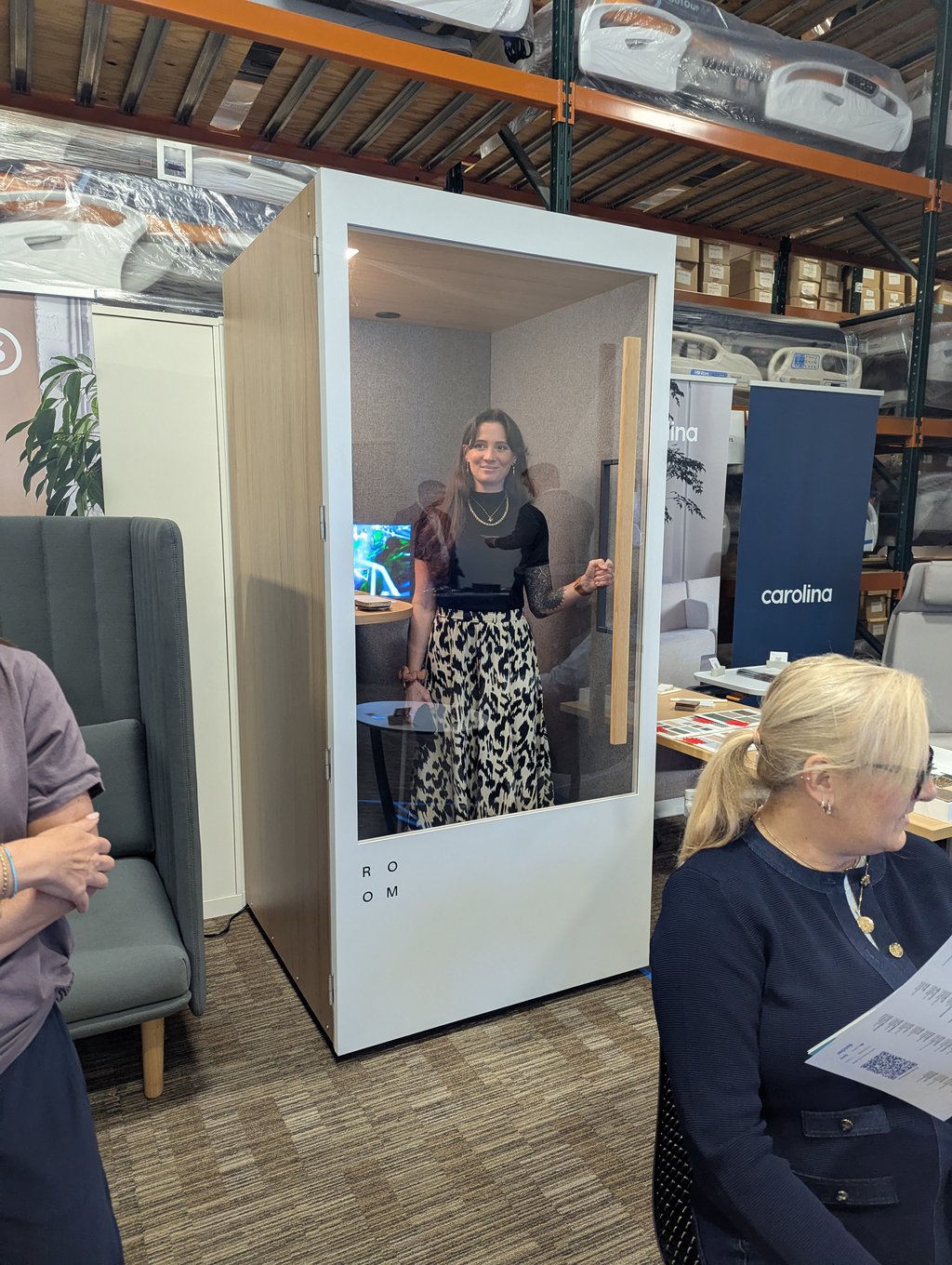 An attendee standing inside a white office pod with a glass door beside the Carolina banner