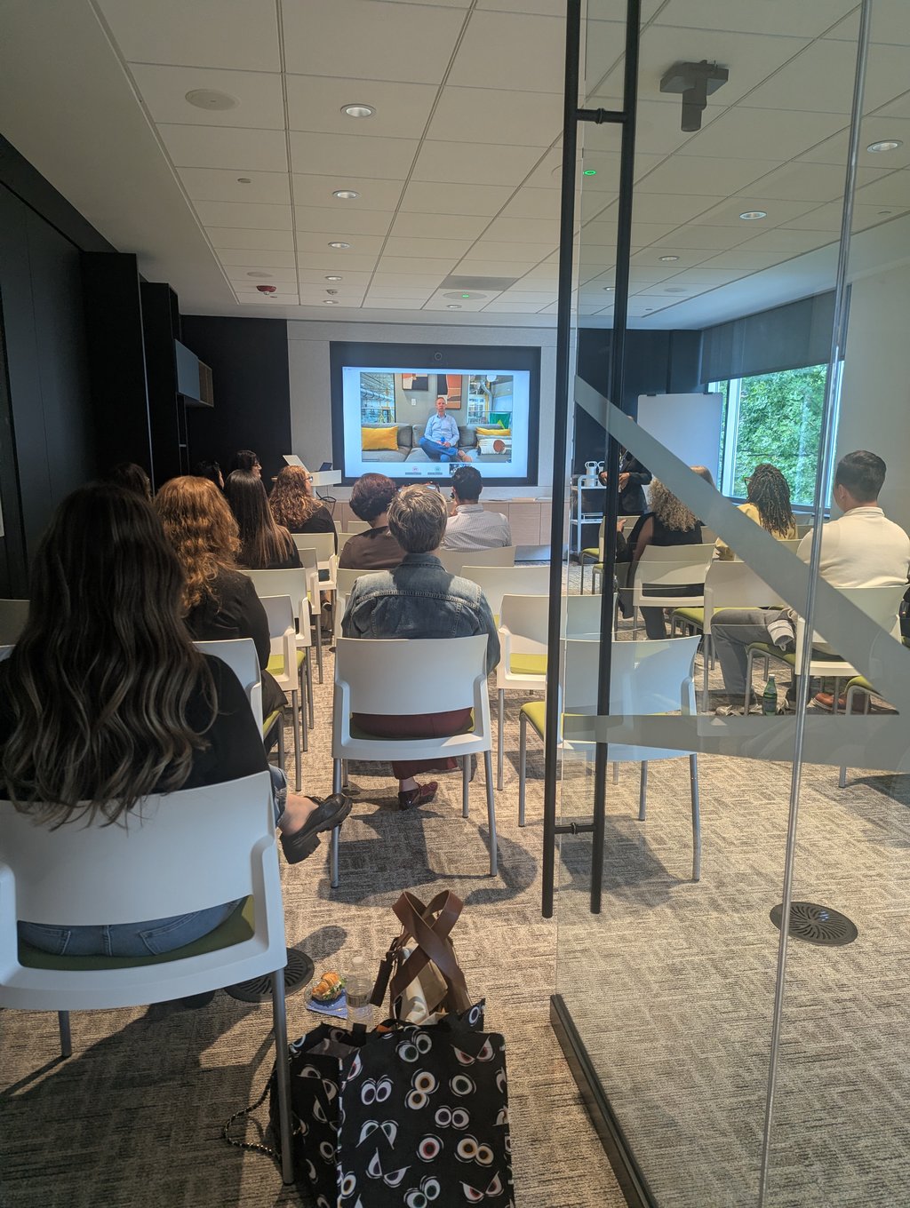 Rows of seated attendees viewed through a glass partition, facing a wall screen showing a video call