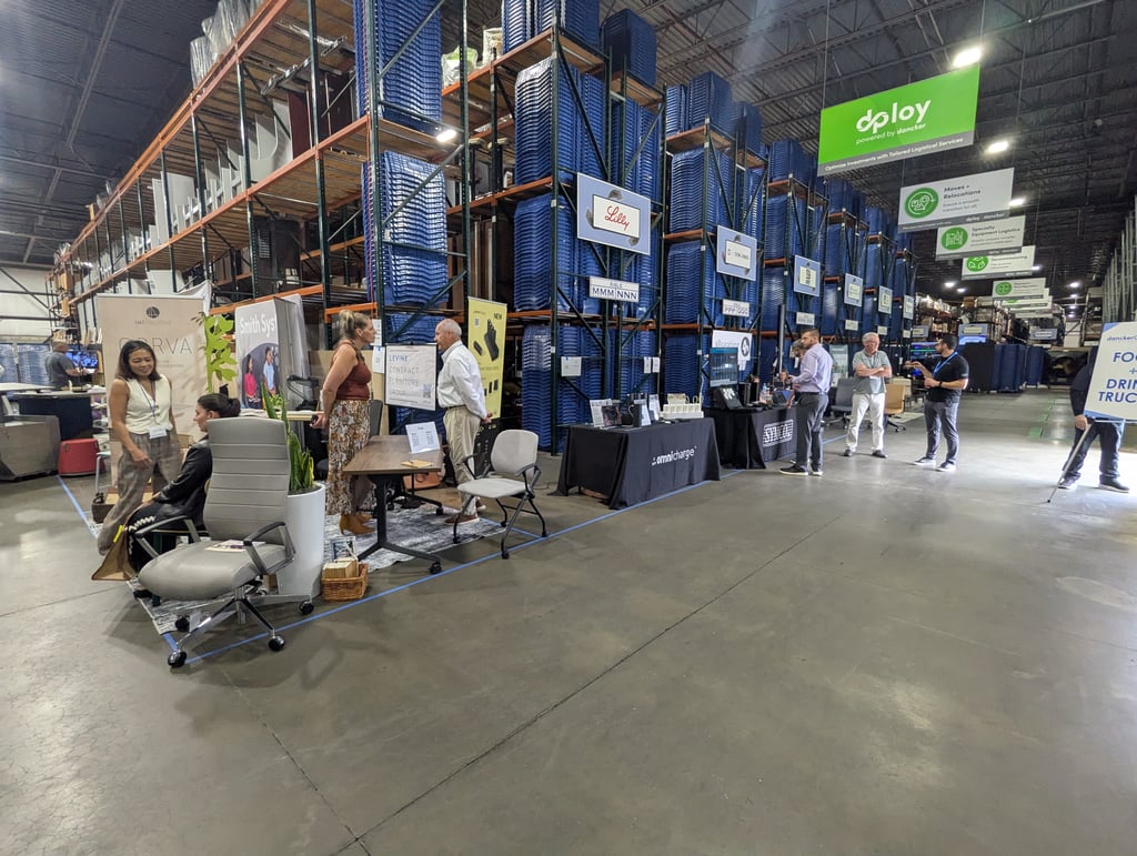 Booths and attendees along an aisle of blue bin racking beneath the green dploy sign