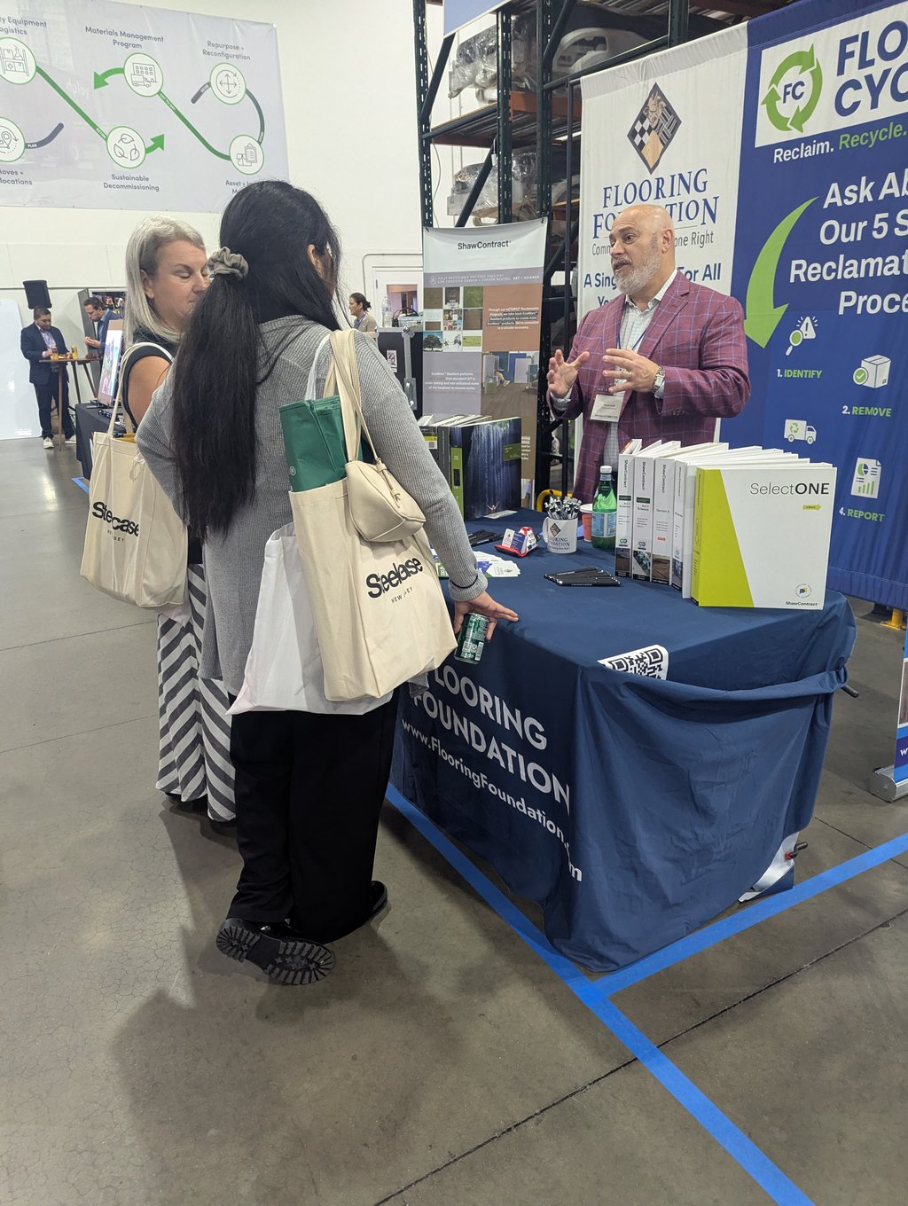 An attendee in a plaid jacket talking with two exhibitors at the Flooring Foundation and ShawContract stand