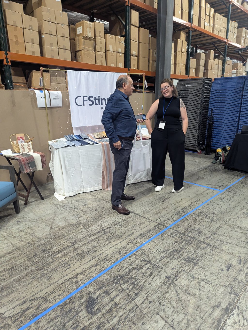Two attendees talking over a table of folded textiles at the CF Stinson booth, number 17