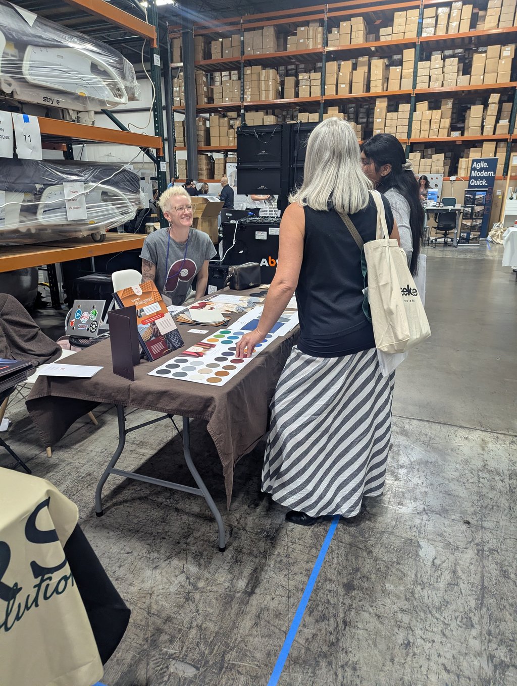 A smiling exhibitor at a table of colour swatches talking with two visitors among the warehouse shelving