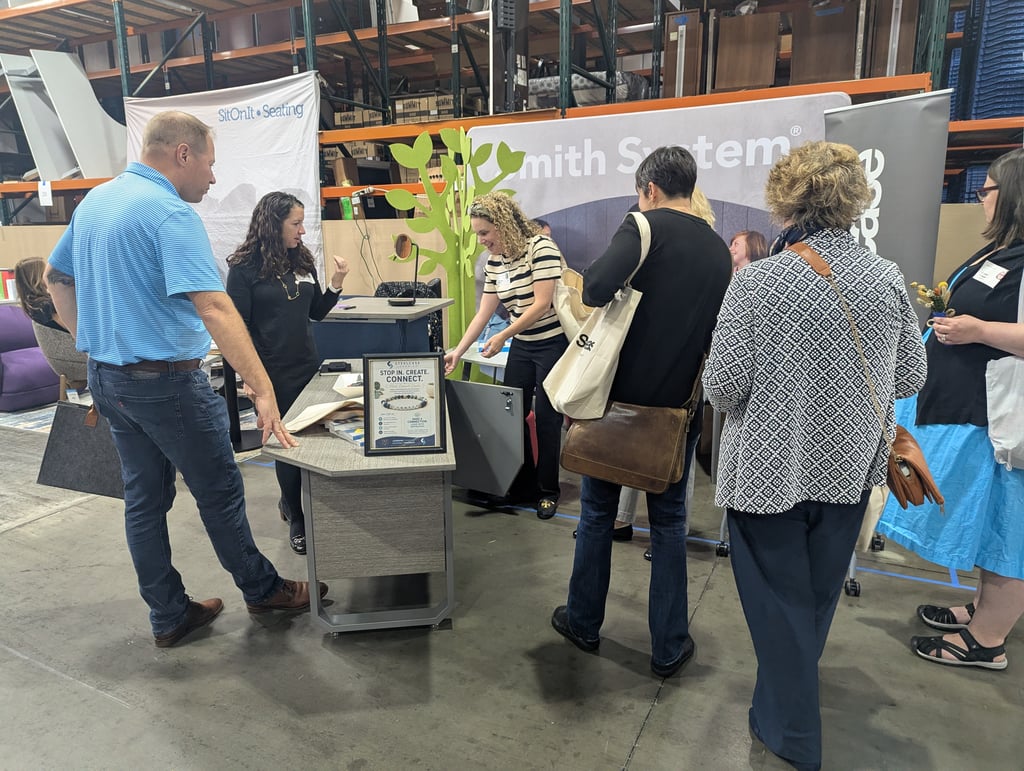 A group of attendees around the SitOnIt Seating demo table with its Stop In Create Connect sign
