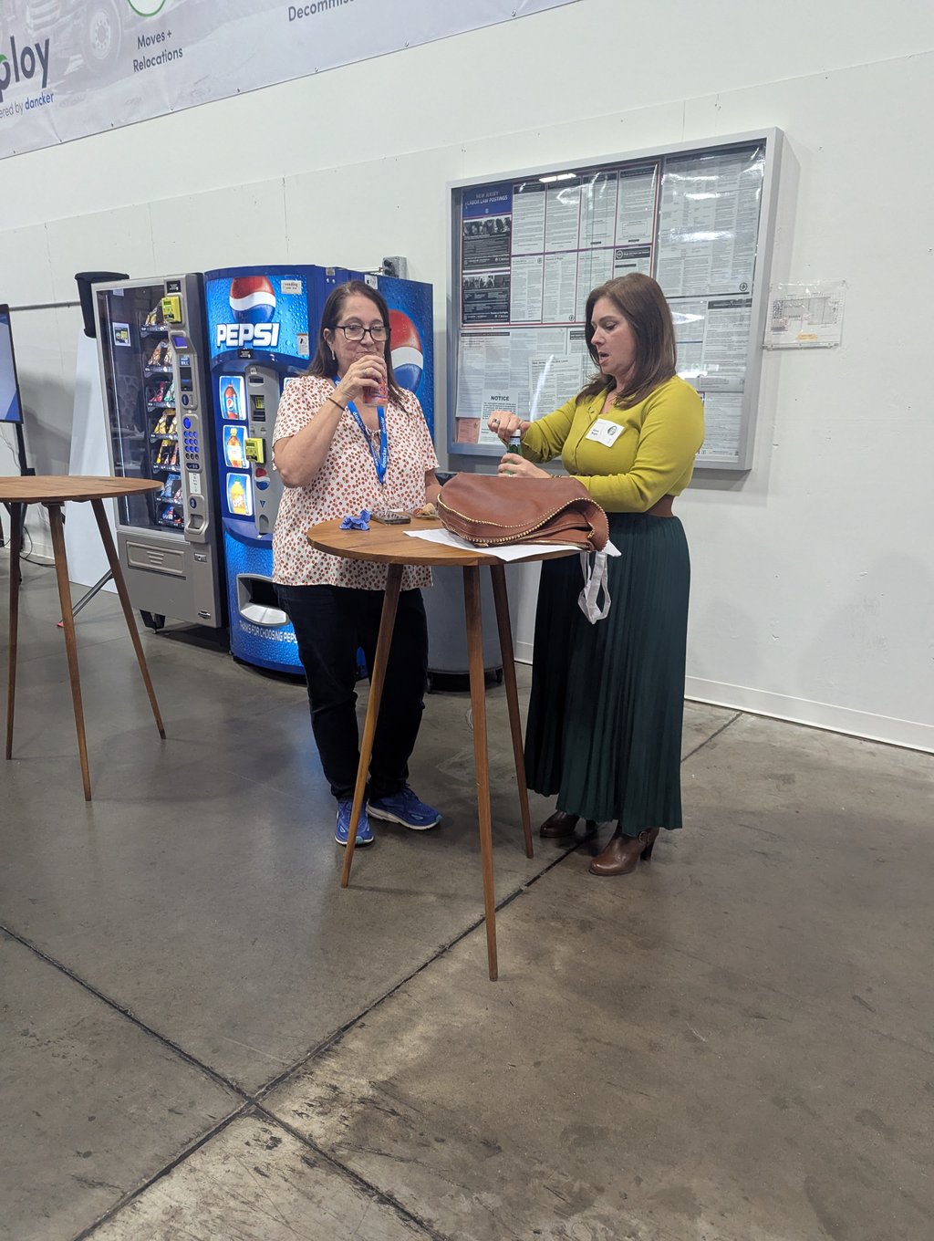 Two attendees with drinks at a tall round table in the break area beside a vending machine