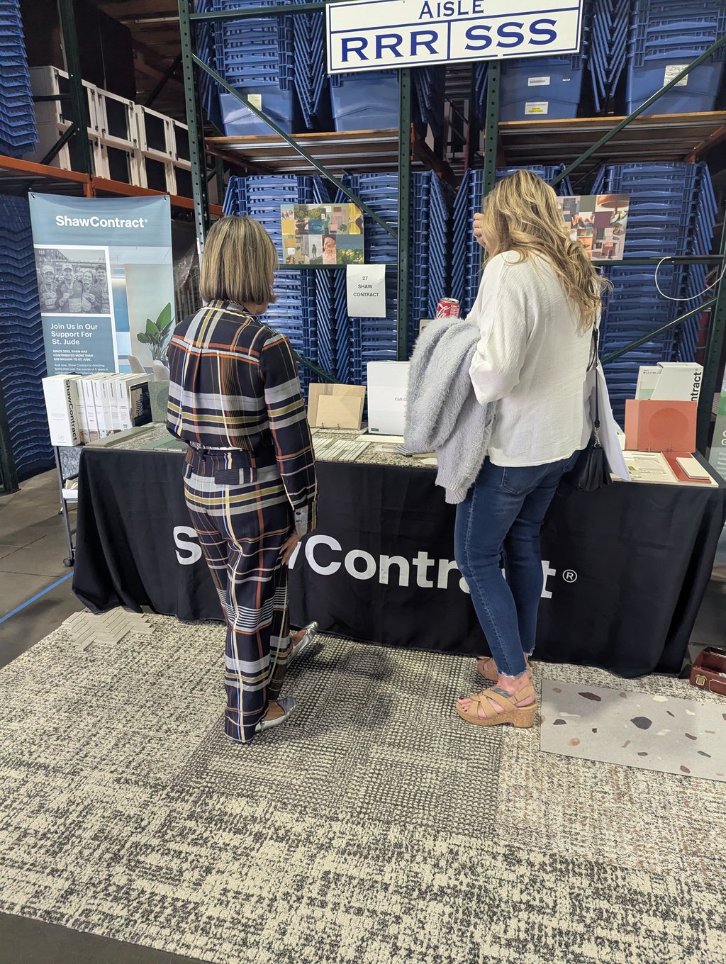 Two attendees examining samples at the ShawContract table against racks of stacked blue bins